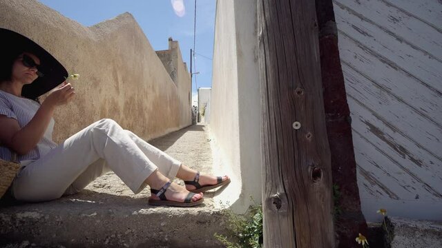 Santorini romantic vacation. Attractive woman sitting on narrow street and smelling fresh spring flower blosom