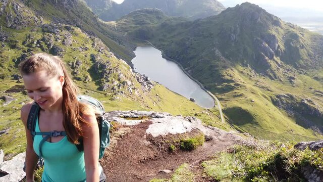 A young female hiker is walking up mountain Mannen near Haukland Beach in Lofoten, Norway. She smiles happily in the sunshine during a warm summer day. Beautiful lake and mountains in the background.