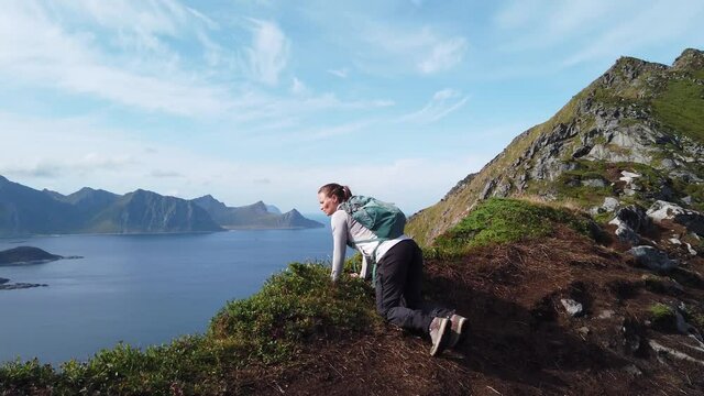 A young woman is hiking up towards the peak of Mannen mountain in Lofoten, Norway near Haukland Beach. She crouches down and sits down right on the edge to look over at the majestic view.