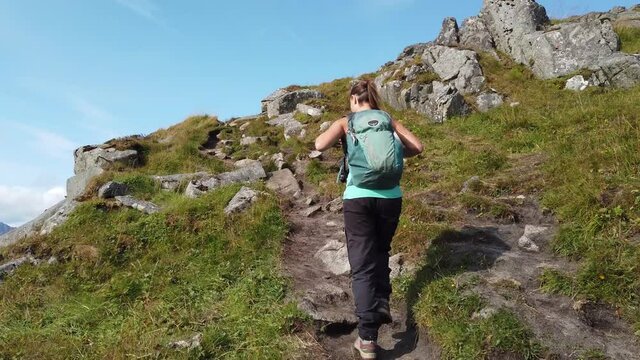 A young brunette woman is walking with her hiking boots and backpack on a trail leading up to the peak of mountain Mannen. On the edge of the cliff leading down to Haukland Beach in Lofoten, Norway.