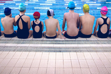Back view of a group of happy young children wearing swimwear sitting at the swimming pool