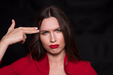 Obraz premium Emotional head shot portrait of a brunette caucasian woman in red dress and with red lips on black background. She make unhappy, dusgusting emotions