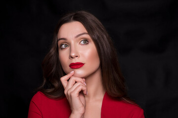 Emotional head shot portrait of a brunette caucasian woman in red dress and with red lips on black background. She thoughtful