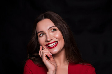 Emotional head shot portrait of a brunette caucasian woman in red dress and with red lips on black background. She smiling with hands near her face