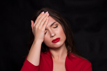 Emotional head shot portrait of a brunette caucasian woman in red dress and with red lips on black background. She have a headache, with fingers on her head