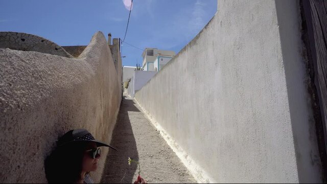 Traveler woman making break sitting on stairs of narrow street in Santorini and smelling spring flower
