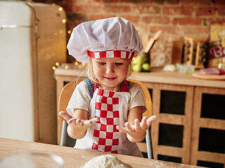 Little girl in cooking chef clothes enjoys kneading the dough. Girls at the kitchen. Family housekeeping.