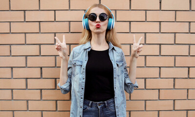 Attractive woman in wireless headphones listening to music blowing red lips sending sweet air kiss wearing a black round hat, jeans jacket over brick wall background