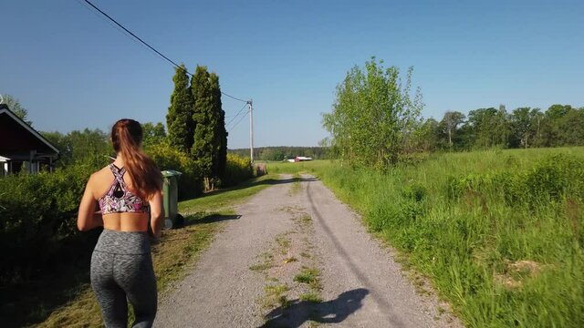 A young, fit and attractive woman is running in leggings and a gym bra top on the countryside in Sweden next to some idyllic and typical Swedish houses in red and white color. Small gravel road.