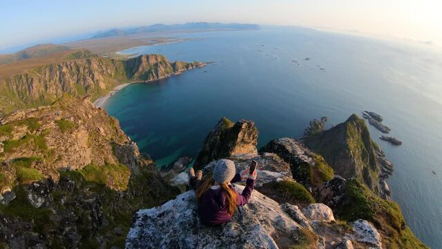 A young woman sits by herself on the edge of a steep cliff on Måtinden mountain in Vesterålen, Norway. She uses her smartphone to take photos of the vast, still, quiet and scenic view in front of her.