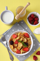 Trendy tiny pancakes for breakfast with strawberry, blackberry and honey in white bowl on yellow background. Closeup