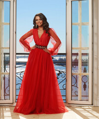 Young beautiful stunning girl in long red dress posing in interior with open door and view on sunny...