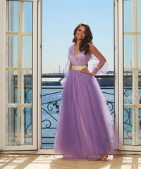 Young beautiful stunning girl in long purple dress posing in interior with open door and view on sunny blue sky. Woman in elegant ball gown
