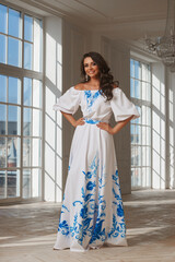 Young beautiful woman with long brown wavy hair in white evening dress standing and posing in...