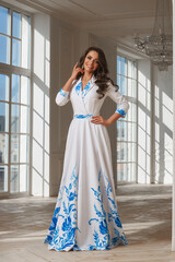 Young beautiful woman with long brown wavy hair in white evening dress standing and posing in...