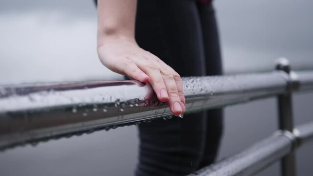 Slow motion female hand erases transparent raindrops from a shiny damp guardrails, drops of water flow on thin fingers.Closeup of caucasian woman touches smooth railing.