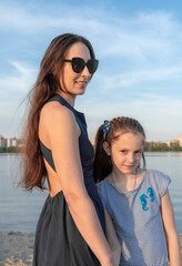 Mother and daughter stand close to each other at the lake shore. The mother smiles, and the girl looks happy. Both are beautiful, of European appearance. High quality photo