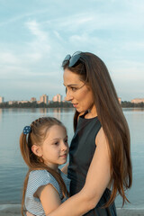 Fototapeta na wymiar Mother and daughter stand close to each other at the lake shore. The mother smiles, and the girl looks happy. Both are beautiful, of European appearance. High quality photo