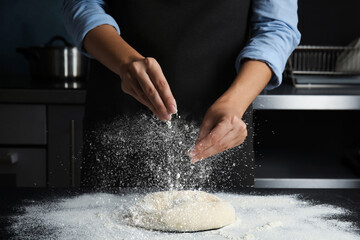 Woman sprinkling flour over dough on table in kitchen, closeup