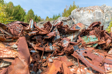 Big pile of scrap metal in an abandoned quarry. The metal is twisted, broken, and rusted. Trees and rock in the background. Closeup view.