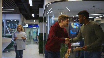 Medium tracking shot of young White and Black men meeting in office corridor, shaking hands, walking together and talking cheerfully