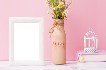 Fragment of an interior with an empty mock up photo frame, a vase with daisies and books