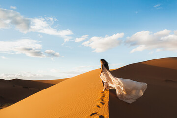 Woman in amazing silk wedding dress with fantastic view of Sahara desert sand dunes in sunset light. Landscape of Morocco, Africa.