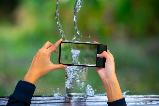 Two Hand Holding Mobile Phone And Take A Photo Drink Water Pouring In To Glass Over Sunlight And Natural Green Background.Water Splash In Glass Select Focus Blurred Background.