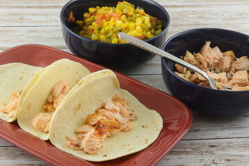Making salmon tacos by putting salmon in tortillas on plate with two filling bowls of baked salmon fillet pieces and mixture of corn, tomatoes, green peppers