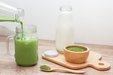 Obraz na płótnie Canvas Pouring iced matcha green tea latte​ from​ buttle into mug with green tea powder and fresh milk in bottle on wooden table
