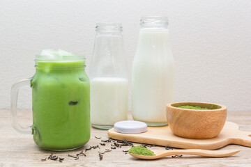 Iced matcha green tea latte in​ mug with green tea powder, dry tea leaves​ and fresh milk in bottle on wooden table