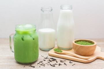 Iced matcha green tea latte in​ mug with green tea powder, dry tea leaves​ and fresh milk in bottle on wooden table