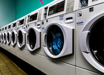 Laundromat line of white washing machines with laundry in them and is empty with teal wall and tile floor.