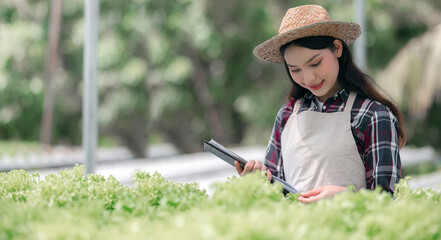 young beautiful woman in plaid shirt, apron and wearing hat holding clipboard while standing in hydroponic vegetable farms.