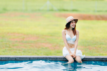 Beautiful Asian woman wear white swim suit with hat sit on the edge of the pool.