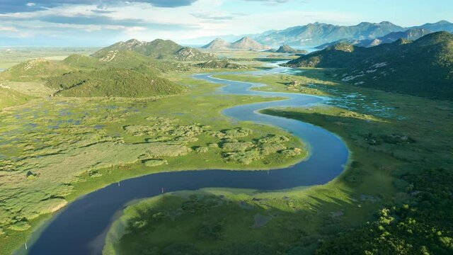 Blue river running through green valley toward distant mountains. Bends and curves of Crnojevica river in Montenegro, meandering through the marsh, on its way to lake Skadar. Aerial footage.