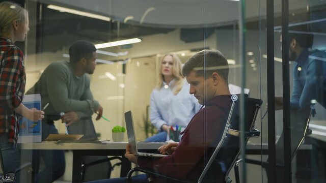 Tracking shot of diverse group of five business people brainstorming in office behind glass wall. Middle aged woman presenting her ideas to colleagues, focus on man using laptop