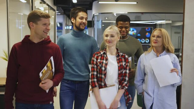 Medium shot of confident business team of five diverse people walking down office corridor and talking joyfully