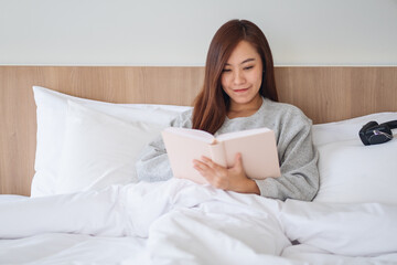 Closeup image of a beautiful young woman reading book in a white cozy bed at home