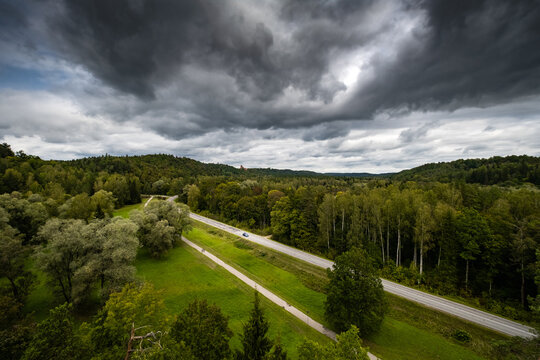 A road in between the meadow and forest under a grey sky with thick cloud with highlight on a car