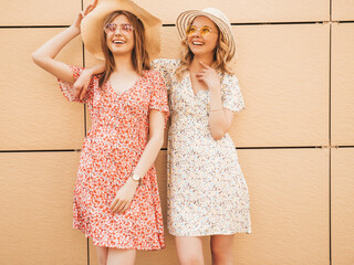 Two young beautiful smiling hipster girls in trendy summer sundress.Sexy carefree women posing near wall on street background in hats. Positive models having fun and hugging