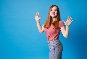 Young ginger girl in pink t-shirt and jeans posing isolated on blue background. Girl raised her hands, admiring gesture.