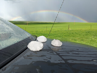 Giant hailstones displayed on a black car after a powerful storm has passed. A rainbow can be seen...