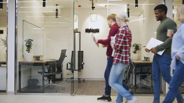 Wide shot of four diverse people entering small office with glass walls, sitting down at desks, talking and starting work