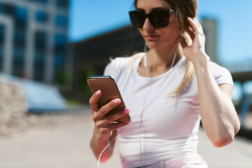 Young fitness woman in sportswear resting after a workout on the promenade sitting at yoga mat and using a smartphone. Healthy lifestyle and sports, street training, social distancing