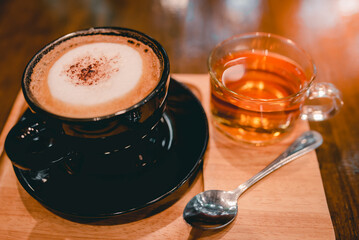 Hot Cappuccino Coffee in a Cup of Black Fresh Coffee With a cup of tea beside each other Beautiful places in a coffee shop The background is a brown wooden table.