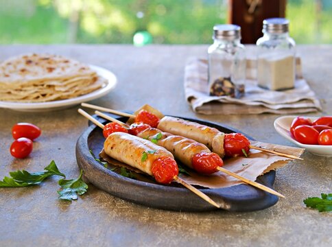 Grilled sausages with tomatoes on skewers on a brown wooden plate