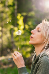 Blonde woman blowing dandelion blossom fluffs in the background of blurred dense green forest during summer evening time and illuminated by sun beams and lens flare