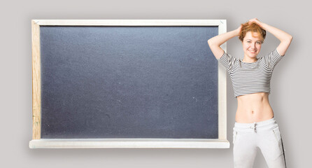 Happy young woman stands at the side of blackboard with copy space and straightens hair. Conceptual slim fit healthy body education.