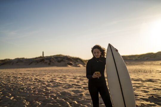 Shallow focus shot of a smiling female looking at her phone while holding a surfboard